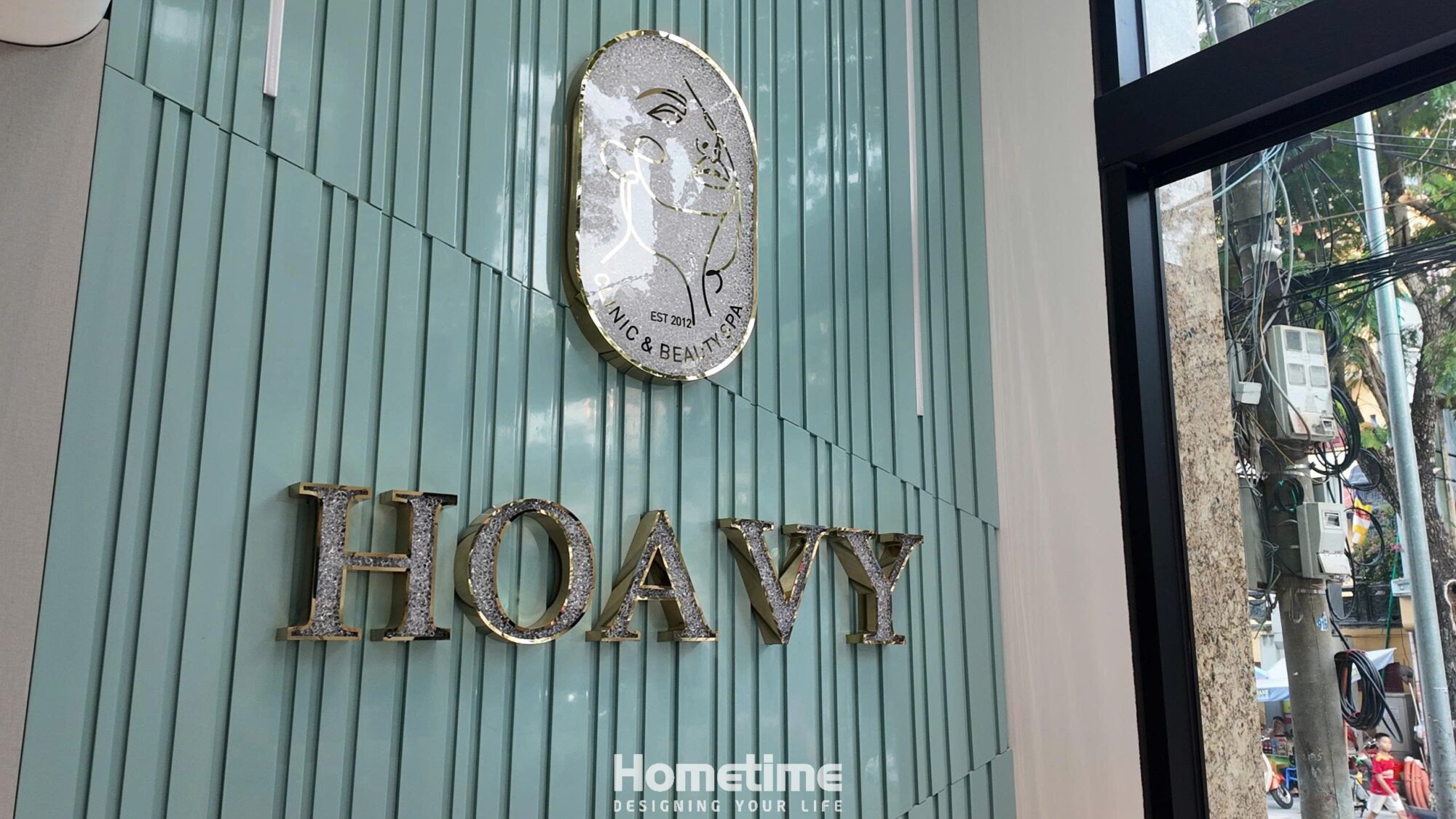Logo Hoa Vy ánh bạc lấp lánh nổi bật trên nền lam sóng xanh dịu, tạo ấn tượng tinh tế và chuyên nghiệp.